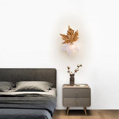 Brass and frosted glass maple leaf sconce glowing on white bedroom wall beside nightstand
