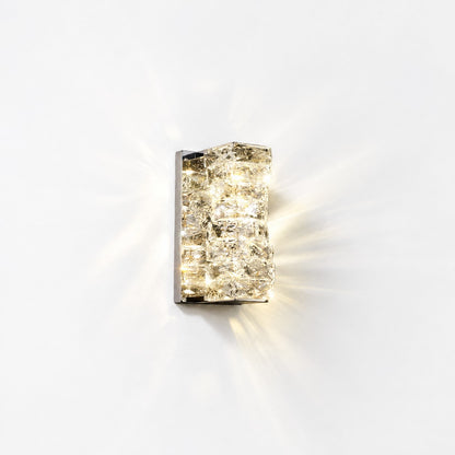 Compact square crystal sconce lit on white wall, warm starburst glow spreading outward